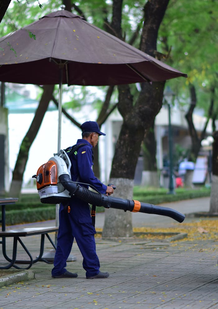 A worker using a leaf blower to clean fallen leaves in an urban park during fall.
